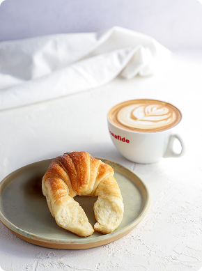 Un croissant dorado se sienta en un plato redondo junto a una taza de capuchino con arte latte en una mesa blanca. Al fondo, un mantel blanco.